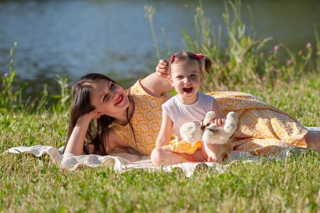 Mother and daughter sitting on a white blanket. Girl holding a white bear. In the background the lake. Horizontally framed shot.の写真素材
