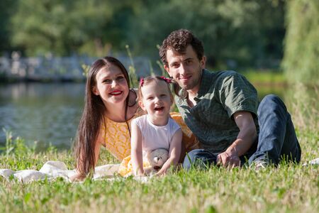 Happy family relaxing in the Park. They are lying on a white plaid. Girl holding in hand a Teddy bear. In the background the lake. Horizontally framed shot.の写真素材
