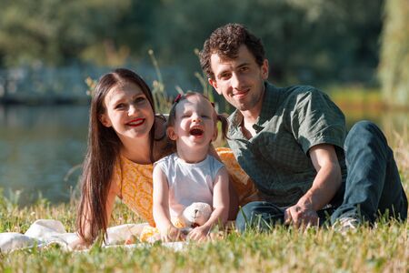 Happy family relaxing in the Park. They are lying on a white plaid. Girl holding in hand a Teddy bear. In the background the lake. Horizontally framed shot.の写真素材