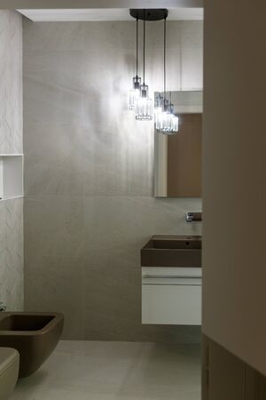 Stylish brown sink, chrome faucet, large mirror and ceiling light three-lamp chandelier. Bidet and toilet. Modern bathroom design. Vertically framed shot.の写真素材
