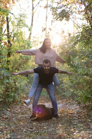 Young beautiful couple and their labrador in the Park. A man holds a woman on his back, and they spread their arms. Rays of setting sun break through the branches of trees. Vertically framed shot.の写真素材