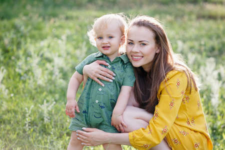 Happy family leisure outdoors. Portrait of smiling young woman holding little toddler son in arms. They are sitting on the green grass. Horizontally framed shot.の写真素材