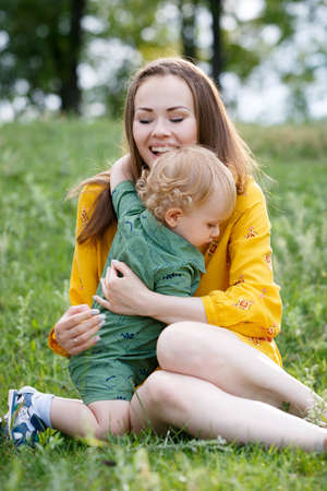 Happy family leisure outdoors. Portrait of smiling young woman holding little toddler son in arms. They are sitting on the green grass. Vertically framed shot.の写真素材