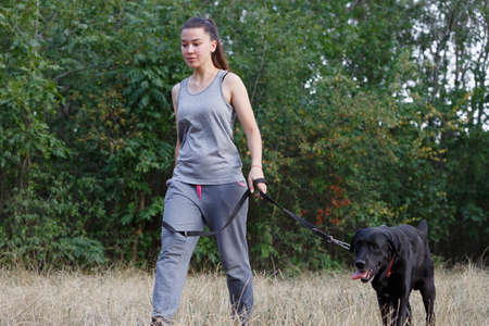 Young attractive sport girl walking with black Labrador in park. Healthy lifestyle. Walk with favorite pet in wood. Horizontally framed shot.の写真素材