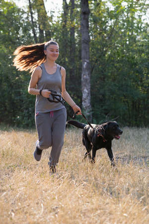 Young attractive sport girl running with black Labrador in park. Healthy lifestyle. Walk with favorite pet in wood. Vertically framed shot.の写真素材