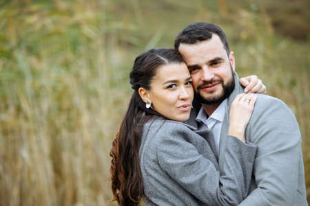 Stylish couple in gray coats gently hugging on the background of reeds. Happy sensual wedding couple embracing. Romantic moments of newlyweds. Horizontally framed shot.の写真素材