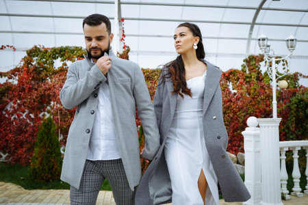 Stylish couple in gray coats walking in the autumn park. Happy sensual wedding couple. Romantic moments of newlyweds. Horizontally framed shot.の写真素材