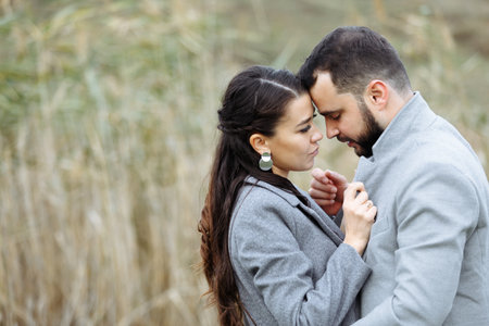 Stylish couple in gray coats gently hugging on the background of reeds. Happy sensual wedding couple embracing. Romantic moments of newlyweds. Horizontally framed shot.の写真素材