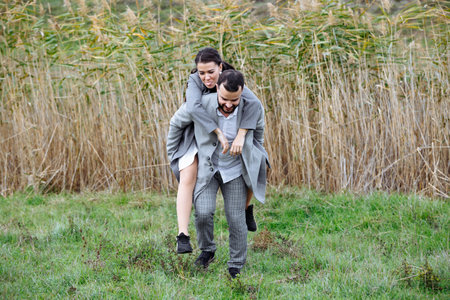 A stylish couple in gray coats are having fun. A man holds a woman on his back and she hugs him. Horizontally framed shot.の写真素材