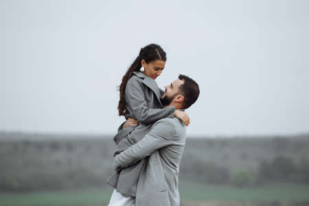 Stylish couple in gray coats on the background of nature. Groom lifts bride up. Happy moments of the wedding day. Horizontally framed shot.の写真素材