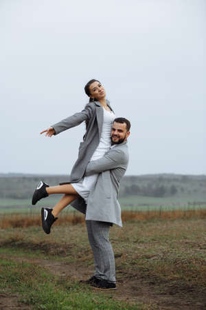 Stylish couple in gray coats on the background of nature. Groom lifts bride up. Happy moments of the wedding day. Vertically framed shot.の写真素材