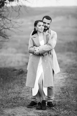 Stylish couple in gray coats gently hugging on the background of nature. Happy sensual wedding couple embracing. Romantic moments of newlyweds. Vertically framed shot.の写真素材