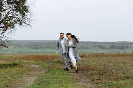 A mischievous couple of newlyweds are running around and having fun in the countryside. Happy moments of the wedding day. Horizontally framed shot.の写真素材