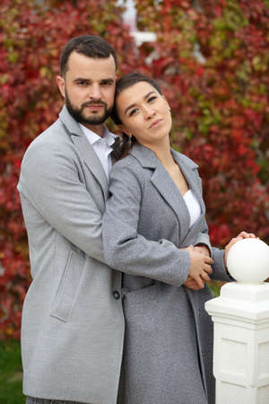 Stylish couple in gray coats gently hugging on the background of autumn leaves. Happy sensual wedding couple embracing. Romantic moments of newlyweds. Vertically framed shot.の写真素材