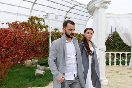 Stylish couple in gray coats walking in the autumn park. Happy sensual wedding couple. Romantic moments of newlyweds. Horizontally framed shot.の写真素材