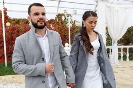 Stylish couple in gray coats walking in the autumn park. Happy sensual wedding couple. Romantic moments of newlyweds. Horizontally framed shot.の写真素材