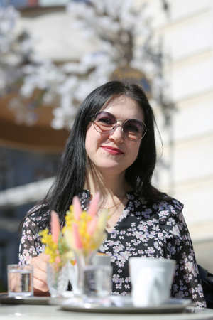 Smiling stylish woman in black shirt with sunglasses sitting at the table drinking coffee in the terrace of restaurant. Vertically framed shot.の写真素材