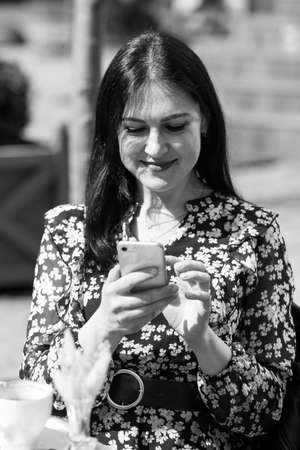 Charming young woman using her smartphone while having cup of coffee. Breakfast on a summer day on terrace in restaurant. Vertically framed shot.の写真素材