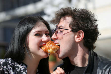 Happy young couple in morning and having breakfast. Cheerful man and woman bite a croissant at the same time. Horizontally framed shot.の写真素材