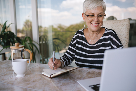 Beautiful senior woman using laptop at home. Mature smiling woman looking at screen while writing in notebook. Portrait of happy modern retired lady with laptop sitting outside on balconyの写真素材