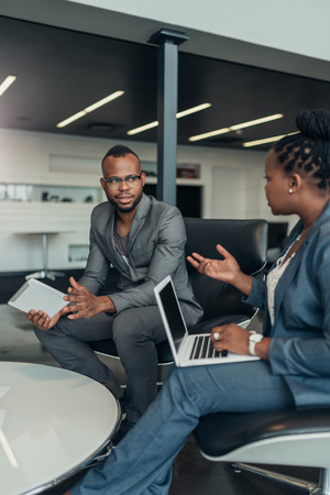 Two african business people talking seriously with hand gestures during a meeting using their laptop and tabletの写真素材