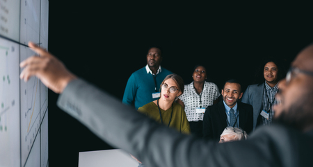 Black african businessman giving a presentation at a business conference. Large video screen with charts and graphs next to himの写真素材