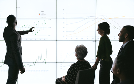 Silhouettes of black african businessman giving a presentation at a business conference to group. Large video screen with charts and graphs in front of him and teamの写真素材