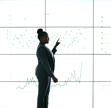 Silhouette of a black african businesswoman giving a presentation at a business conference. Pointing at a large video screen with charts and graphs next to her in a dark roomの写真素材