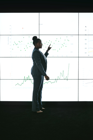 Silhouette of a black african businesswoman giving a presentation at a business conference. Pointing at a large video screen with charts and graphs next to her in a dark roomの写真素材