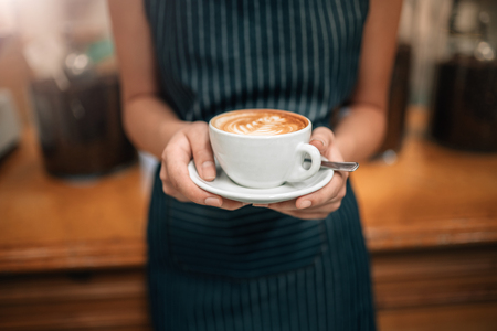 close up of a waitress holding a well poured coffeeCropped Close up of female serving coffee with latte art while standing in coffee shop. Focus on female hands placing a cup of coffeeの写真素材