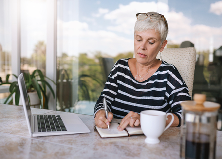 Mature beautiful woman making notes while working on her laptop at home. Elderly woman writing in her notebook outside on the balconyの写真素材