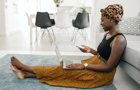 Beautiful African woman sitting on floor in lounge, work call meeting on phone, using laptop at home...