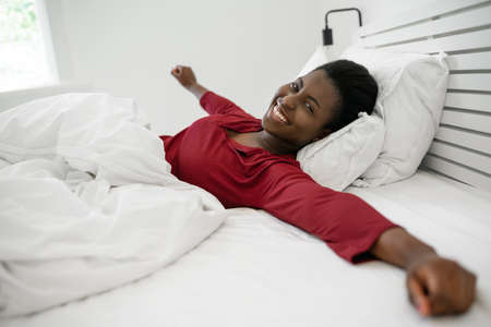 Beautiful African woman waking up in the morning, stretching and smiling, looking into cameraの写真素材