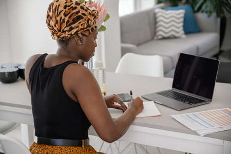 African woman studying from home, looking at laptop, writing down notes. Laptop and smartphone on home office tableの写真素材