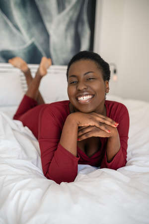Close up portrait of beautiful African woman laying on bed with legs and arms crossed, smiling and looking into the camera with copy spaceの写真素材
