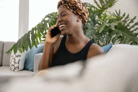 Beautiful traditional young gen z African woman sitting on sofa at home, talking smart phone, smiling, shot with copy spaceの写真素材