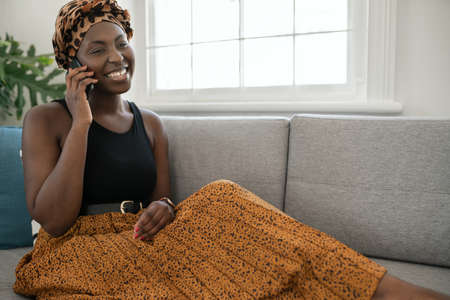 Beautiful traditional young Black gen z African woman sitting on sofa at home, talking smart phone, smiling, shot with copy spaceの写真素材