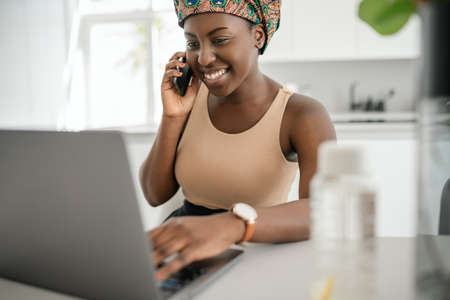 Beautiful young African woman wearing tradition headscarf. Sitting at home working on laptop. On phone call smiling and gesturing with handの写真素材