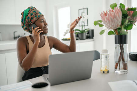 Beautiful young Black African woman wearing tradition headscarf. Sitting at home working on laptop. On phone call smilingの写真素材
