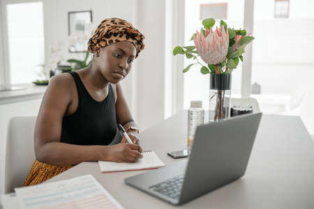 Black African woman working from home, looking at laptop, writing down notes. Laptop and smartphone on home office tableの写真素材