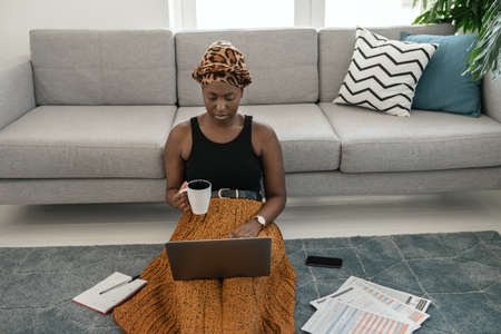 Beautiful black African woman sitting on floor in lounge, studying on laptop at home with coffee in hand wearing traditional head scarfの写真素材