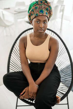Portrait of young fashionable Black African woman. Sitting in modern chair looking seriously into camera wearing traditional African head scarfの写真素材