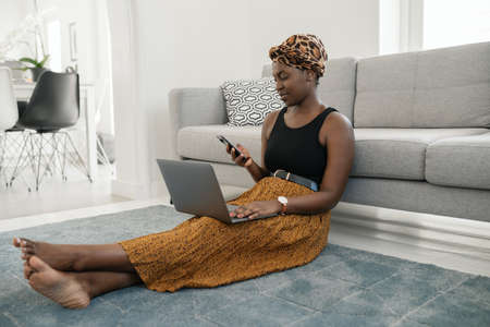 Beautiful black African woman sitting on floor in lounge, work call meeting on phone, using laptop at home wearing traditional head scarfの写真素材
