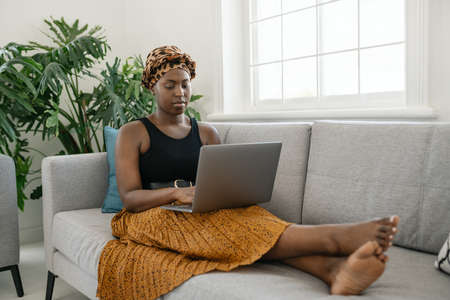 Beautiful traditional young Black gen z African woman sitting on sofa at home, typing on laptop, smiling, shot with copy spaceの写真素材