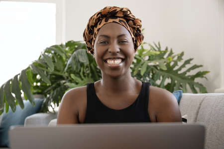 Portrait of beautiful young gen z Black African woman wearing traditional head scarf, sitting in lounge with laptopの写真素材