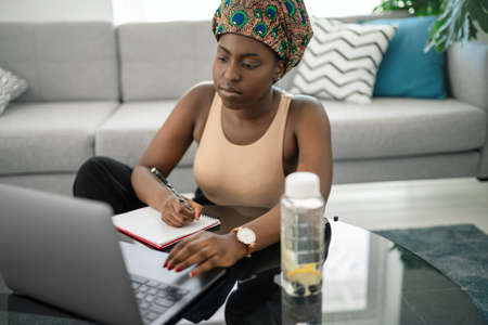 African woman studying from home, looking at laptop, writing down notes. Laptop and smartphone on home office tableの写真素材