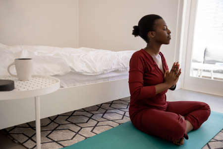 Beautiful Black African woman sitting on yoga mat in bedroom. Meditating and practicing yoga. Early morning exercise. Shot with copy spaceの写真素材
