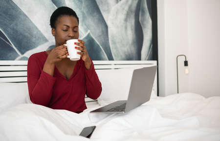 African woman in pajamas with laptop computer and mug sitting in bed and drinking coffee, watching movies or series on her laptopの写真素材