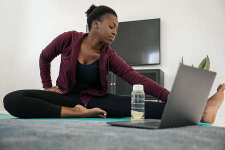 Close up of young Black African woman following online fitness class on laptop, stretching eyes closed, exercise on yoga mat in modern living room at homeの写真素材