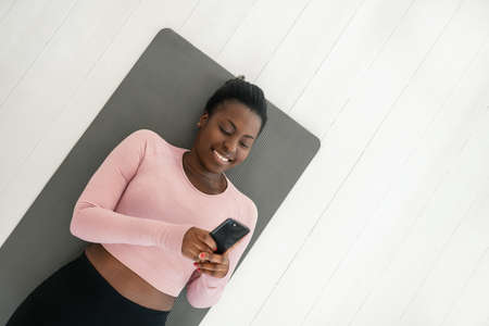 Black African woman laying on yoga mat smiling and looking at phone, resting and taking a break from exercising. shot from above with copy spaceの写真素材